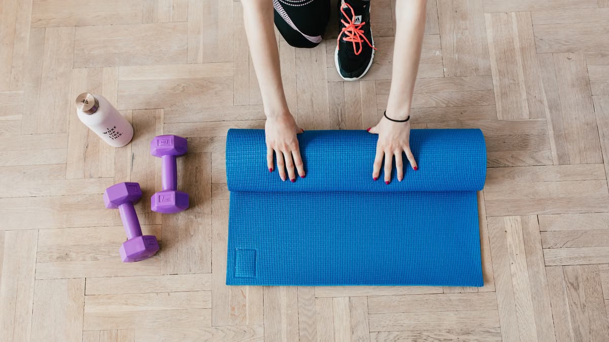 Top view of anonymous female athlete in black leggings and sneakers unfolding blue mat for exercising on floor near dumbbells and water bottle in modern fitness center